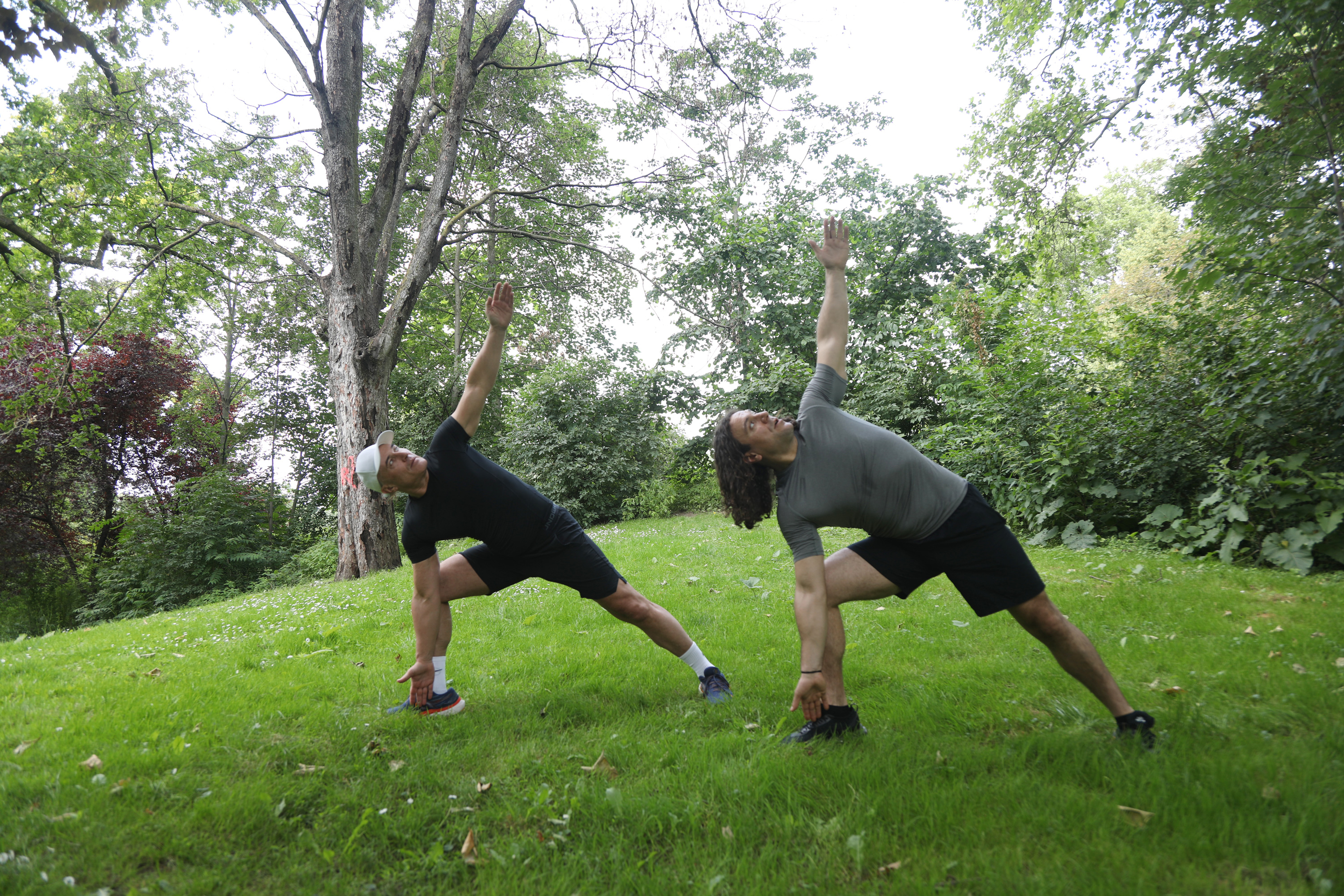 Outdoor Yoga Exercises - Stretching Two men practicing stretching exercises outdoors on a green meadow with trees in the background.