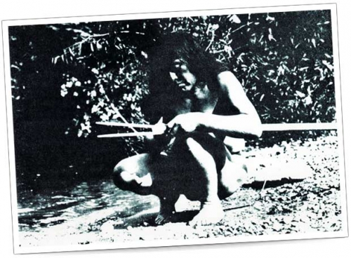 Traditional Hunting Method – Black and White Photo A man sits by a riverbank crafting a spear, barefoot in natural clothing.