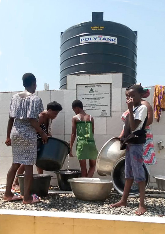Water Tank - Water Extraction in Containers A group of children extracting water from a water tank, various containers, wearing bright clothes in a sunny setting.
