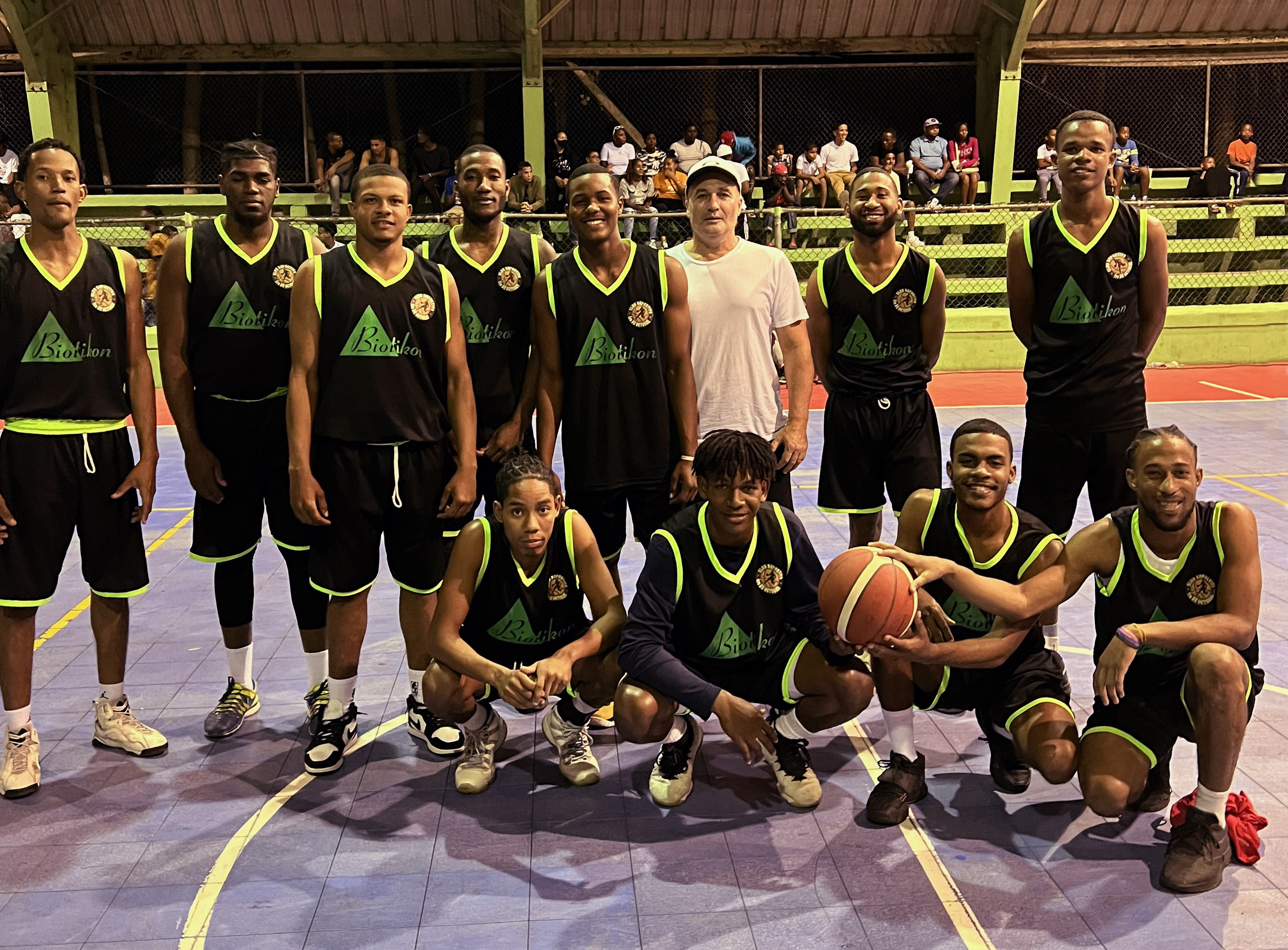 Basketball Team – Group Photo A group of basketball players in black jerseys with green accents, standing on the court, holding a basketball and cheering.